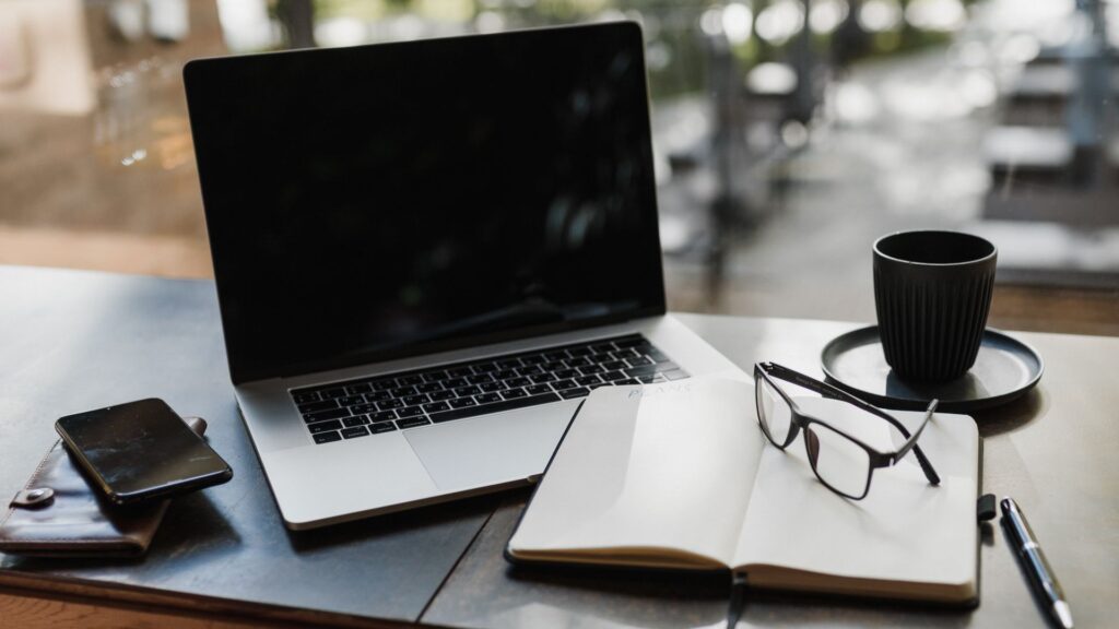 Laptop on desk next to a pair of glasses, notebook, and cup of coffee.