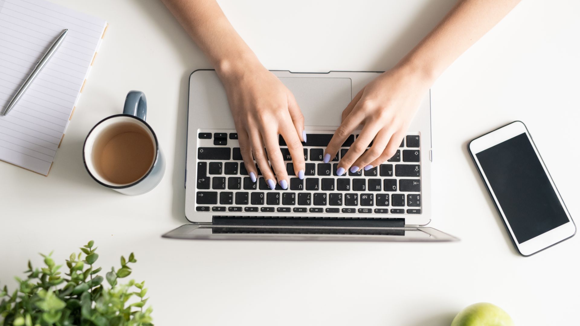 Laptop on desk next to a cup of coffe, smartphone, notepad, plant, and an apple.