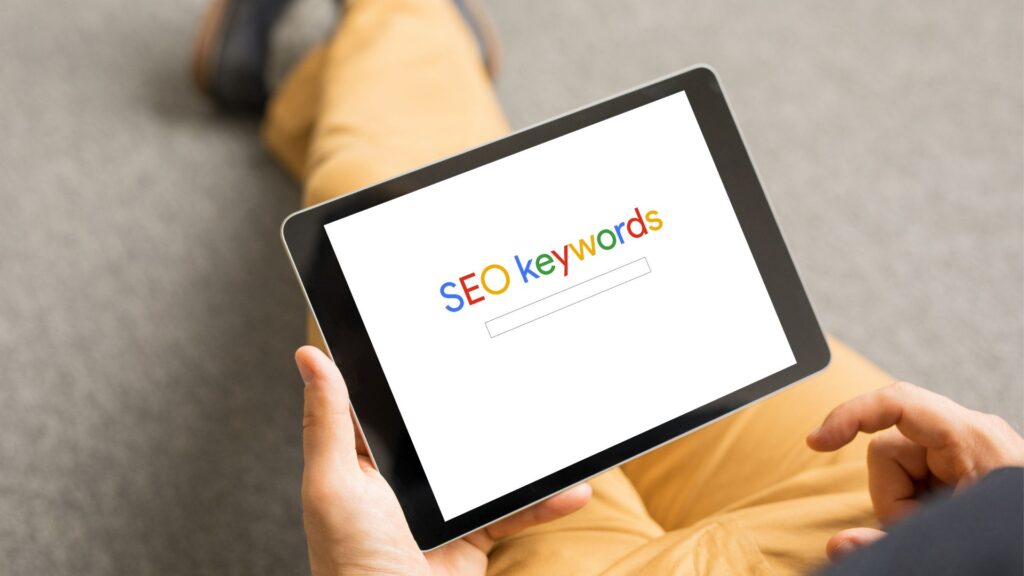 Person sitting on floor holding a tablet with the words "SEO Keywords" on the screen.