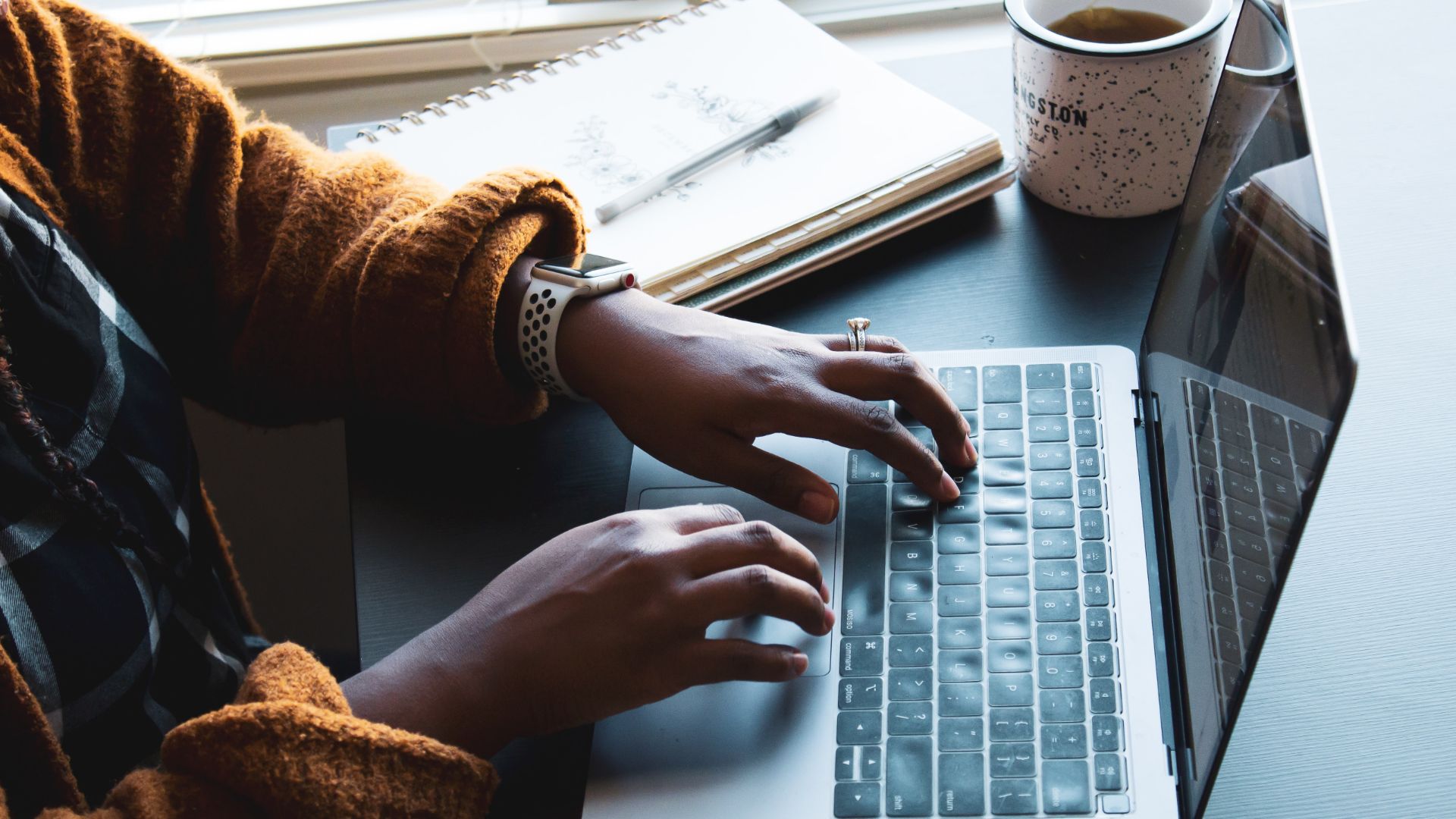 Person with hands on a laptop keyboard on a desk next to a notepad and a cup of coffee.