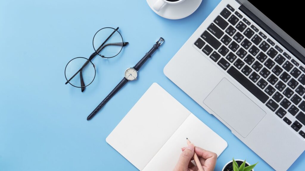 SEO expert writing in notepad with a laptop, a pair of glasses, a clock, and a cup of coffe on the desk.